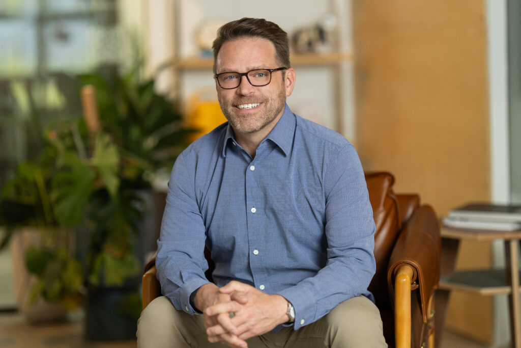 A man in glasses and a blue shirt sits smiling in a cozy room with plants and shelves, creating the perfect headshot for his portfolio.