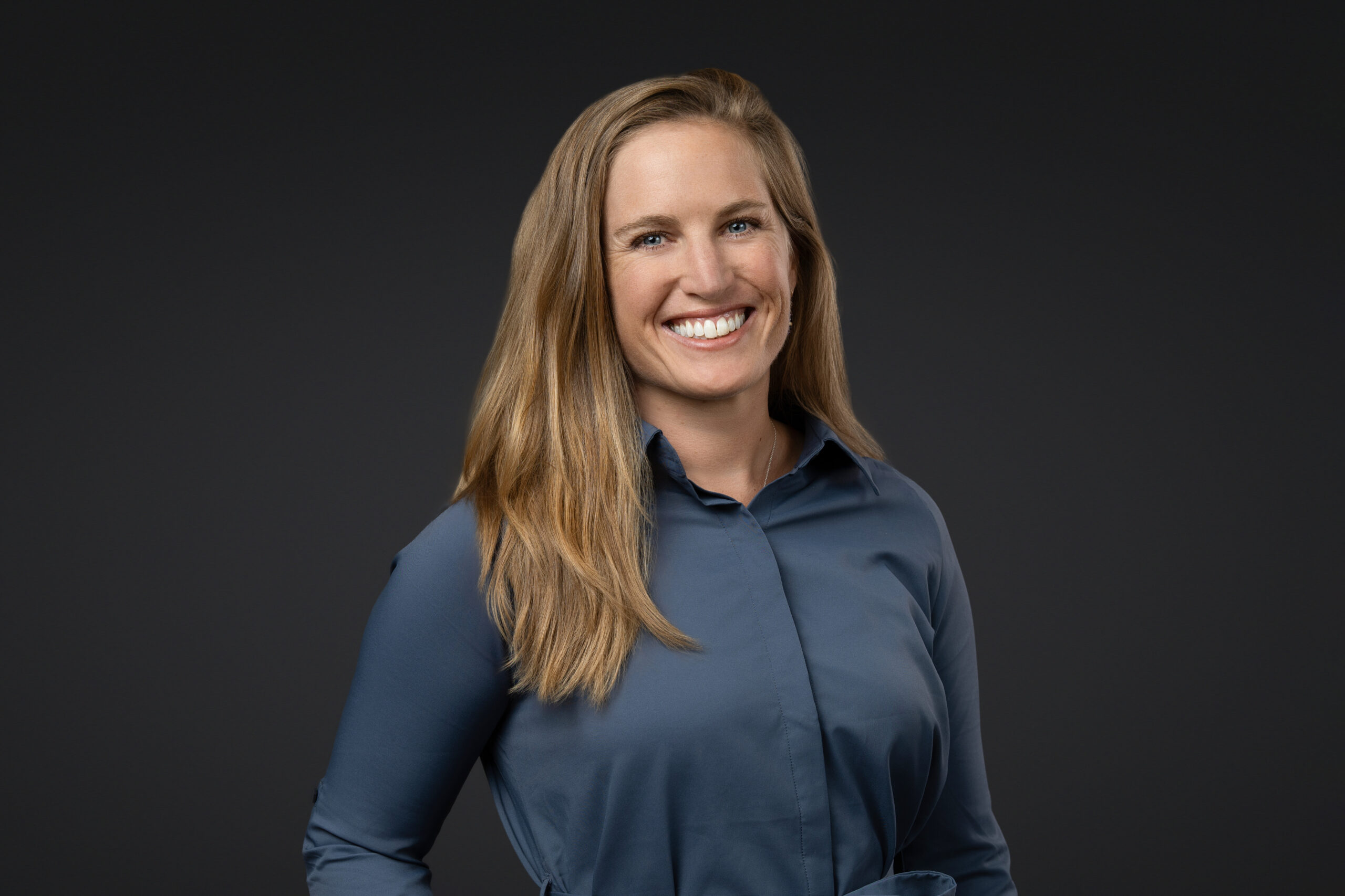 A person with long blonde hair smiles in a headshot, wearing a blue button-up shirt against a dark background.