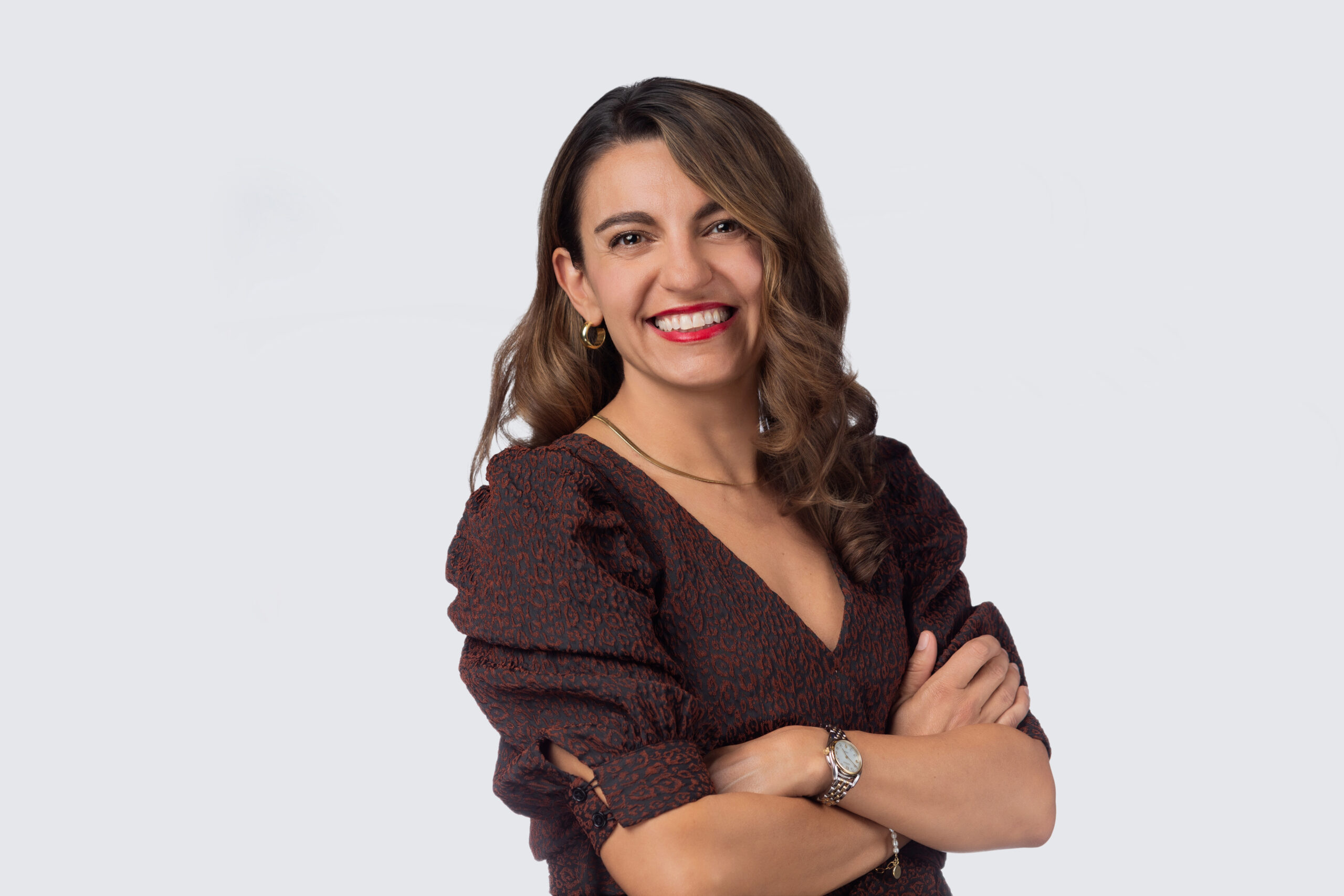 Smiling woman with long brown hair wearing a dark patterned blouse, standing with arms crossed against a plain white background, creating the perfect headshot for her portfolio.