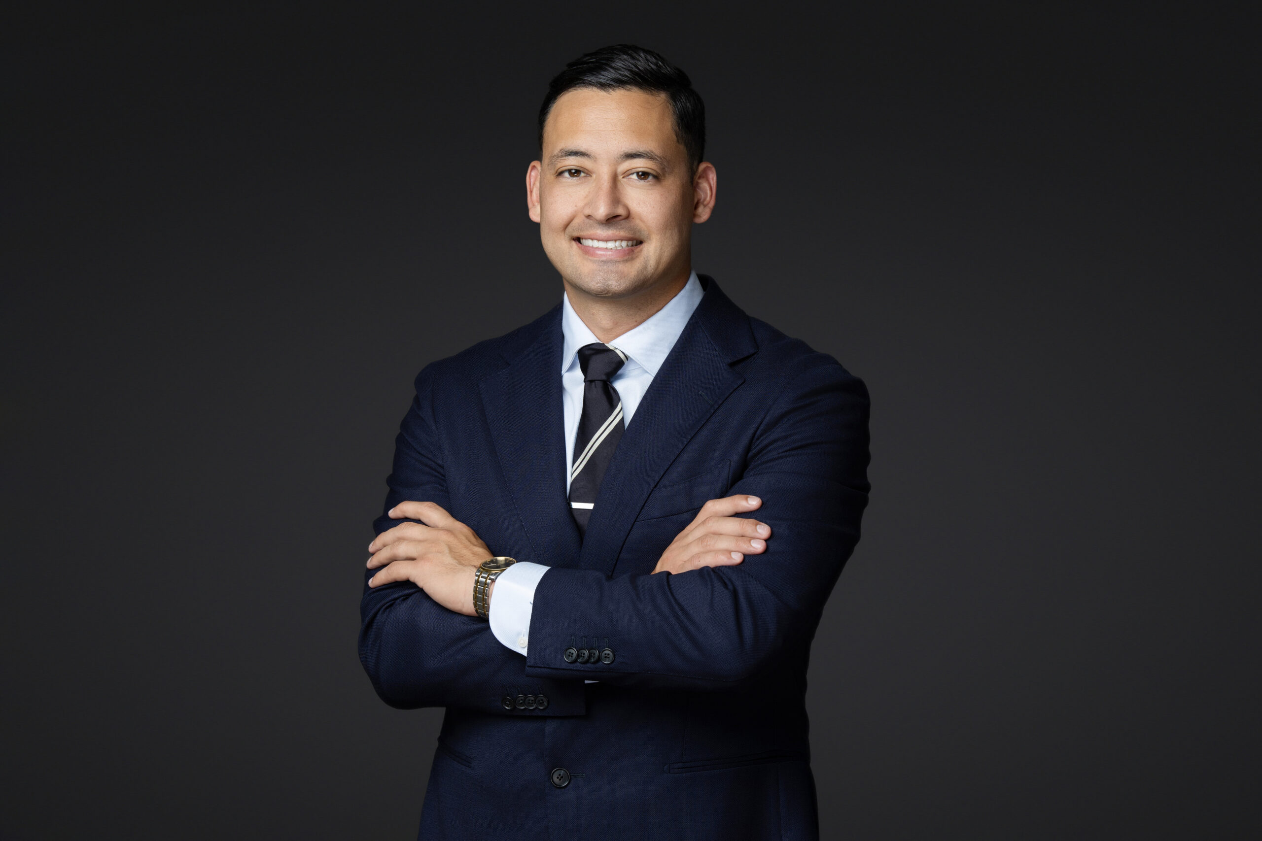 A man in a dark suit and tie stands with arms crossed, smiling confidently for a professional headshot against a plain dark background.