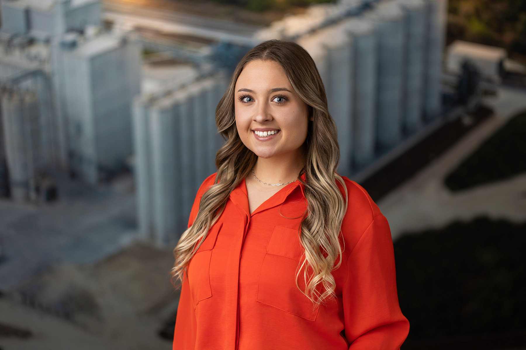 Person in an orange shirt stands smiling for a portfolio headshot, set against a blurred industrial background.