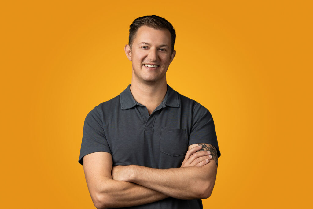 A smiling person with short hair wearing a gray polo shirt poses for a headshot, arms confidently crossed against an orange background, perfect for a portfolio.