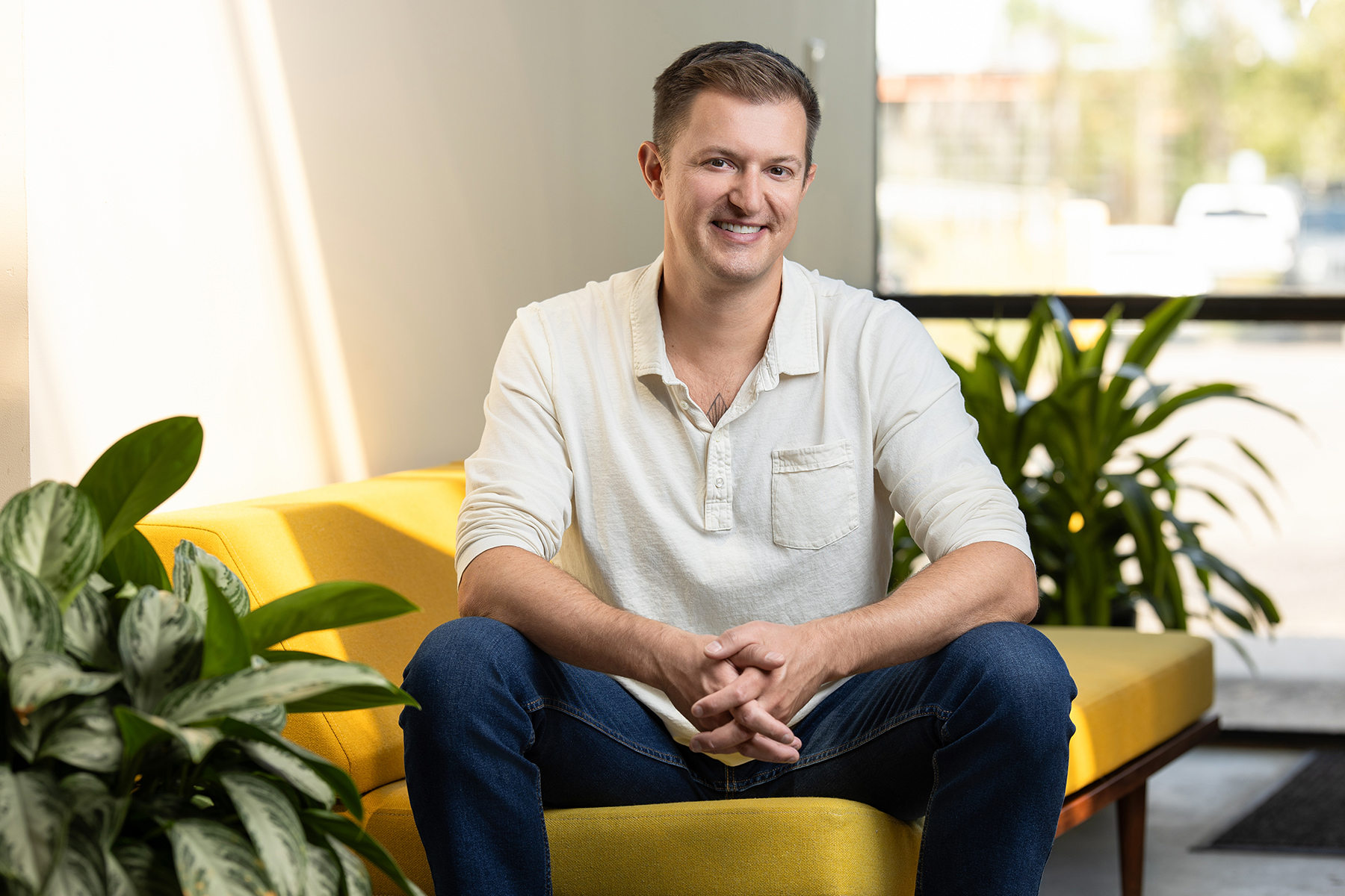 Person sitting on a yellow couch, wearing a white long-sleeve shirt and jeans, with plants nearby—a perfect headshot for their portfolio.