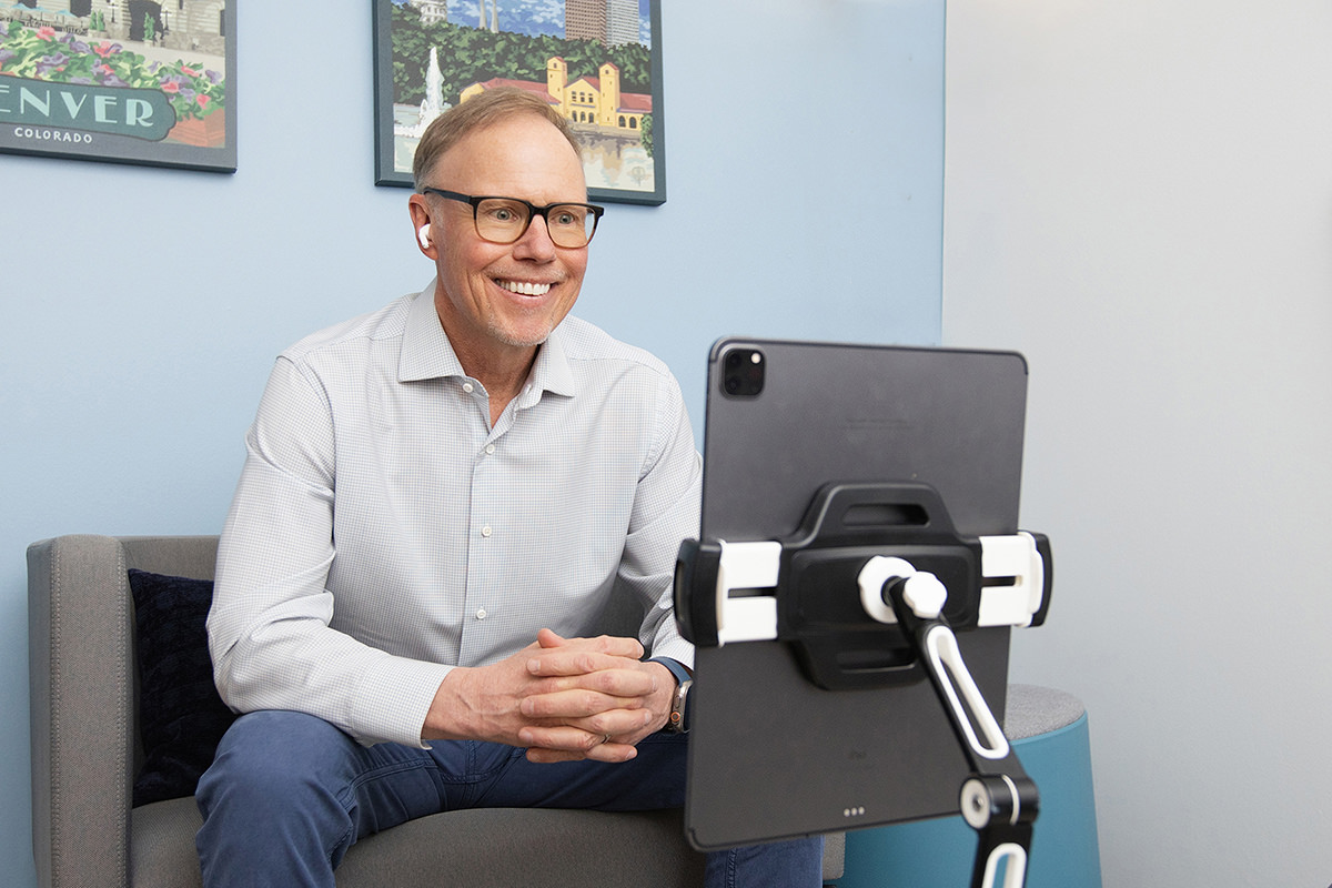 A man in glasses and a button-up shirt smiles while sitting on a couch, using a tablet on a stand for a video call. Behind him, posters of group photography decorate the wall.