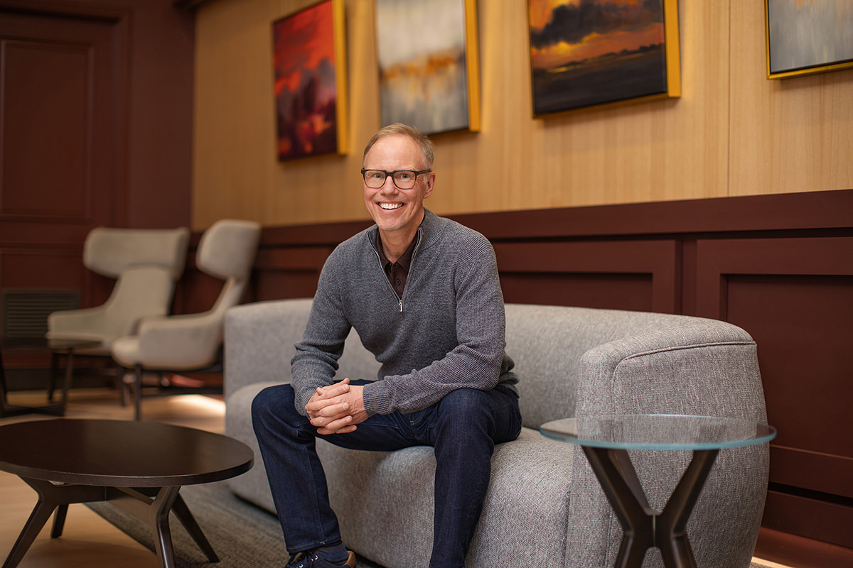 A person is sitting on a gray sofa, smiling warmly in front of a lively group photography scene, with captivating artwork adorning the wall behind them.