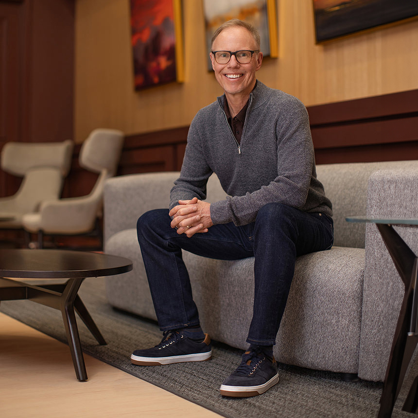 A person in glasses sits on a gray couch, smiling with hands clasped, ready for group photography. They're wearing a cozy gray sweater, jeans, and sneakers.