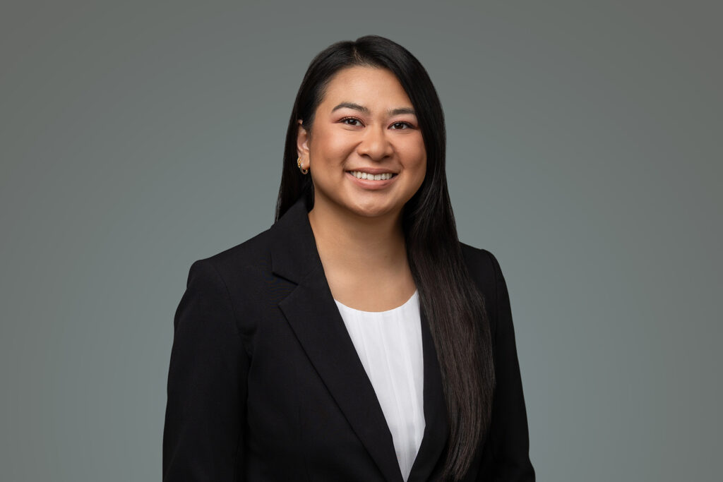 A person with long dark hair wearing a black blazer and white shirt smiles confidently in this professional headshot, set against a plain gray background—perfect for any portfolio.