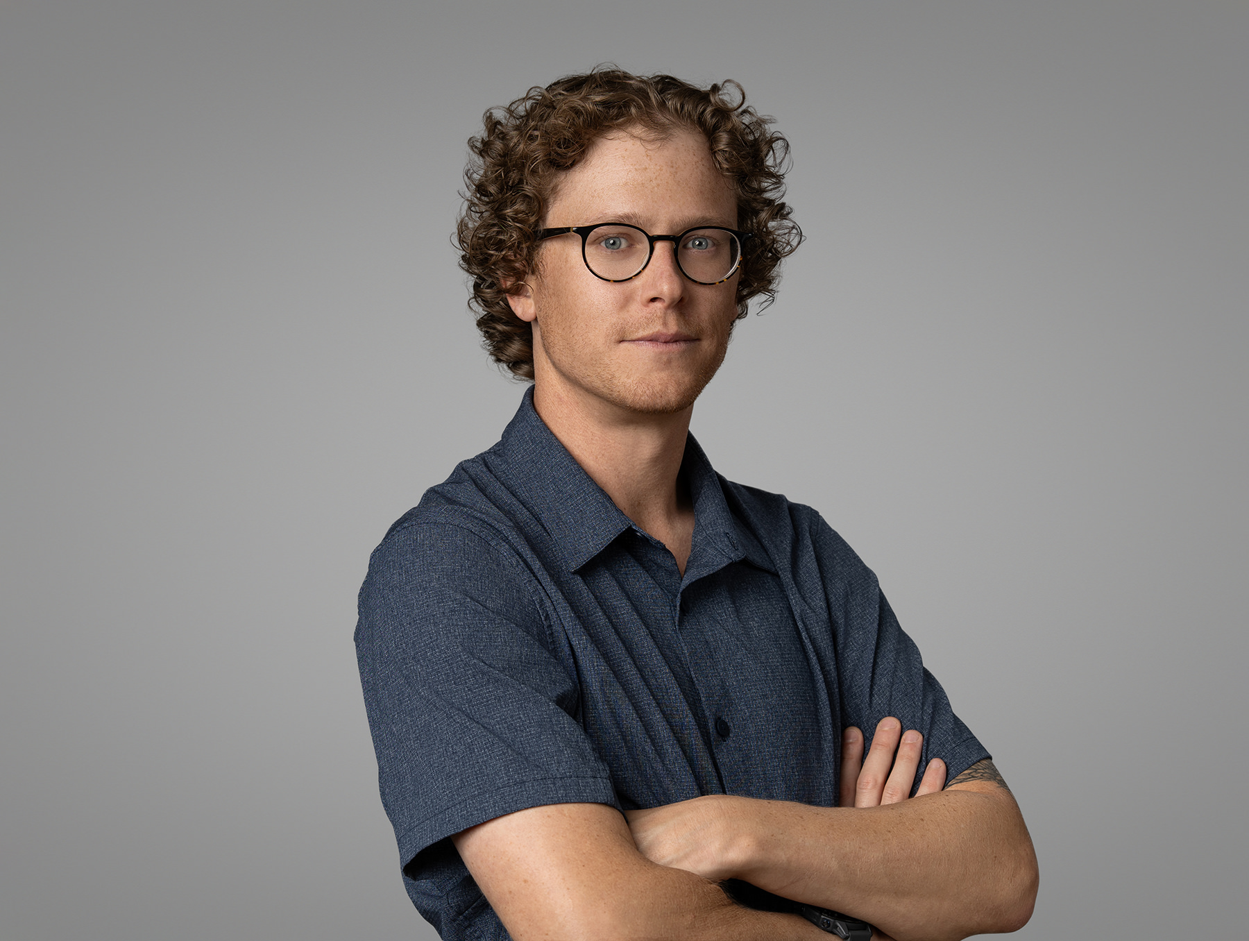 A person with curly hair and glasses stands with arms crossed, wearing a dark blue short-sleeve shirt, creating the perfect headshot for their portfolio against a plain gray background.