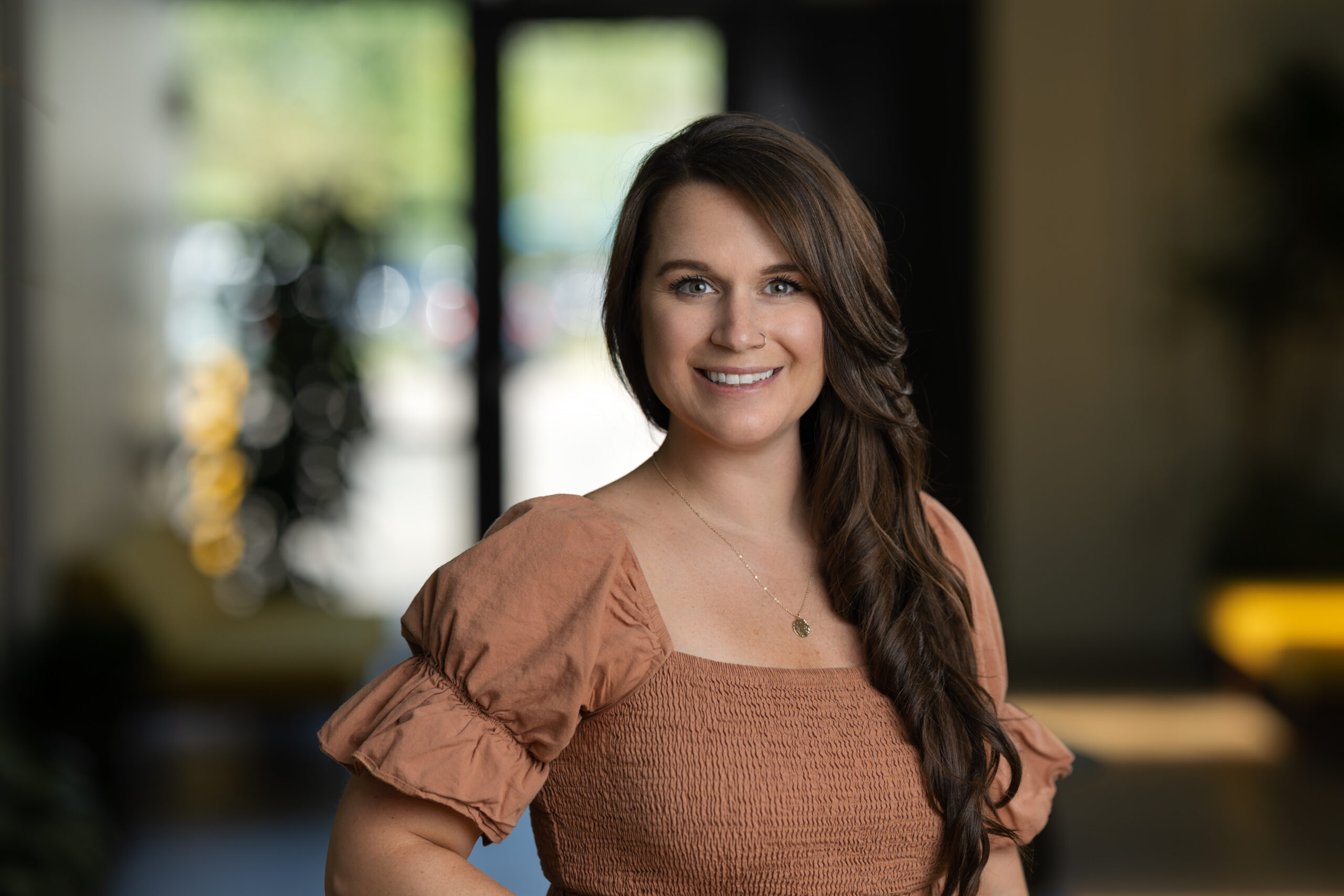 A woman with long hair is smiling in a headshot, wearing a brown shirt with puffed sleeves. The background is softly blurred, capturing her radiant expression perfectly for a portfolio.