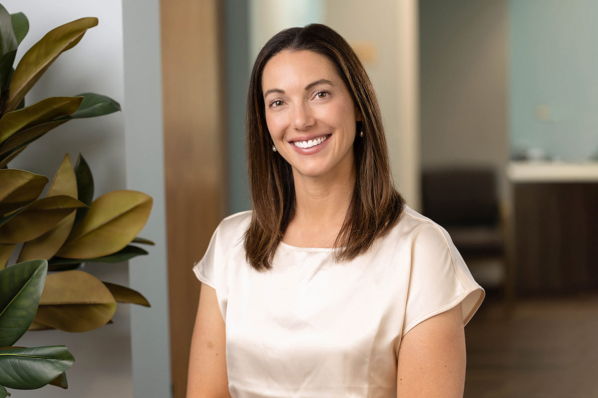 A woman with long brown hair smiles while seated indoors near a plant, subtly bringing warmth and charm to the setting. She wears a short-sleeved white blouse, perfectly complementing the ambiance often sought in group photography.