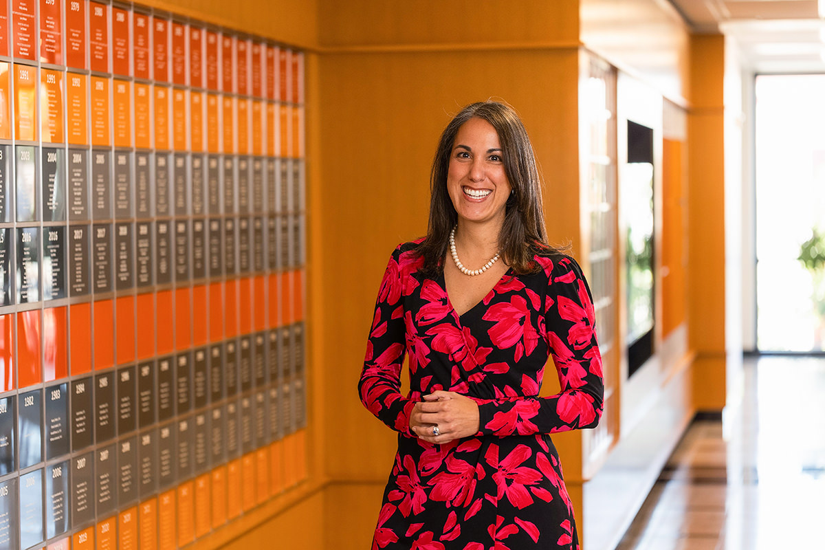 A woman in a red floral dress stands in a hallway with orange and silver plaques on the wall, posed as if waiting for group photography to begin.