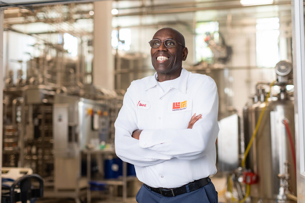 A person in a white uniform with a logo stands confidently among a group, capturing the essence of teamwork in a factory setting with industrial equipment in the background.