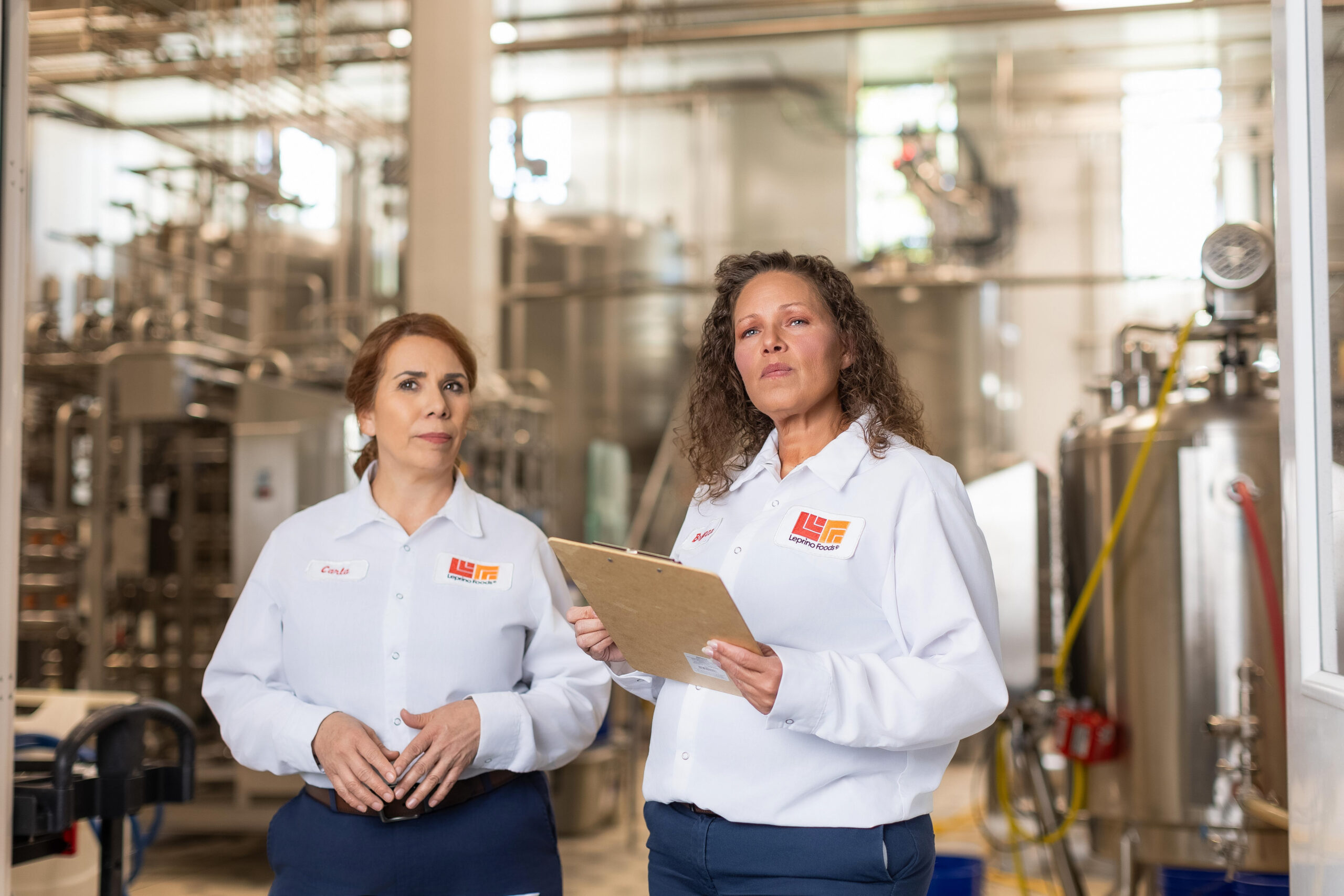 In an industrial setting, two women in white company uniforms stand confidently for a group photography session, one holding a clipboard. Machinery hums in the background, capturing the essence of their collaborative work environment.