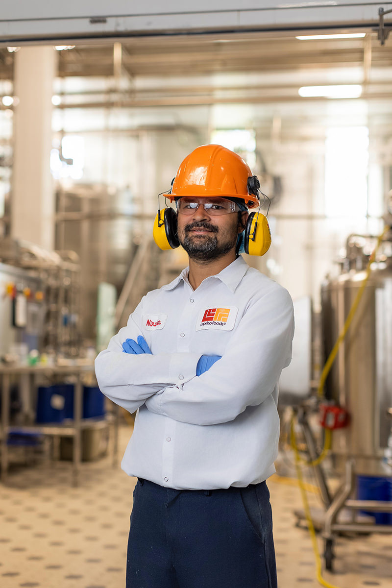 In an industrial setting, a person in safety gear stands confidently with crossed arms, wearing an orange helmet, ear protection, and a white shirt. The scene captures the essence of group photography by emphasizing teamwork and safety in the workplace.