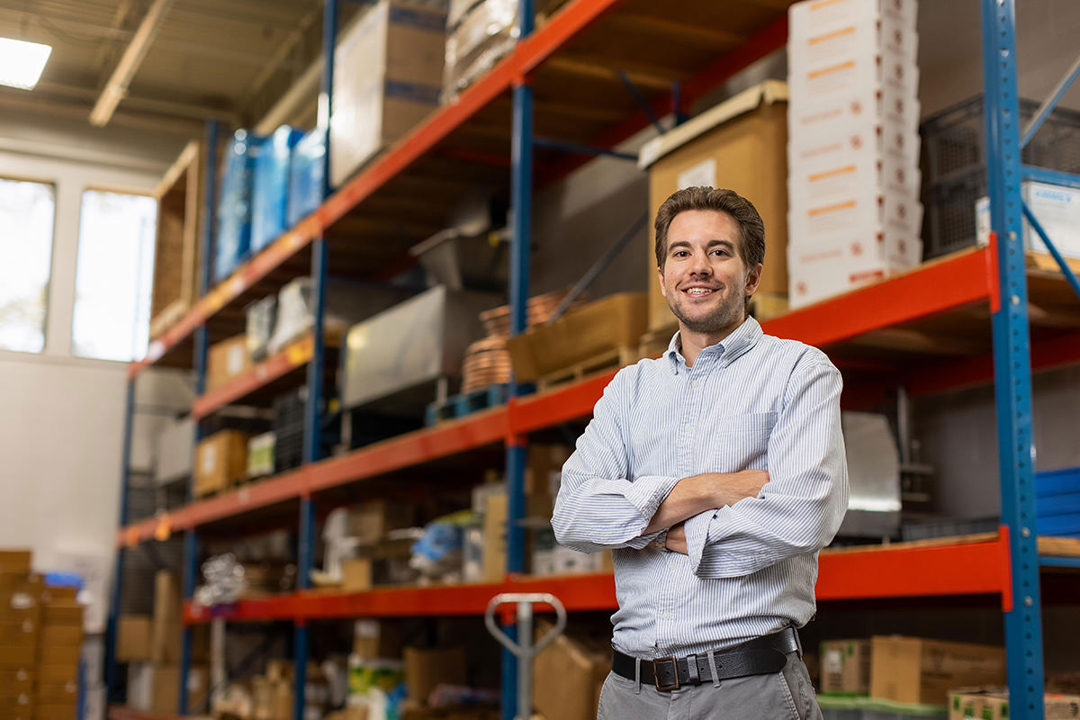 A person stands in a warehouse, smiling with arms crossed, as though waiting for a group photography session in front of shelves filled with boxes and various items.