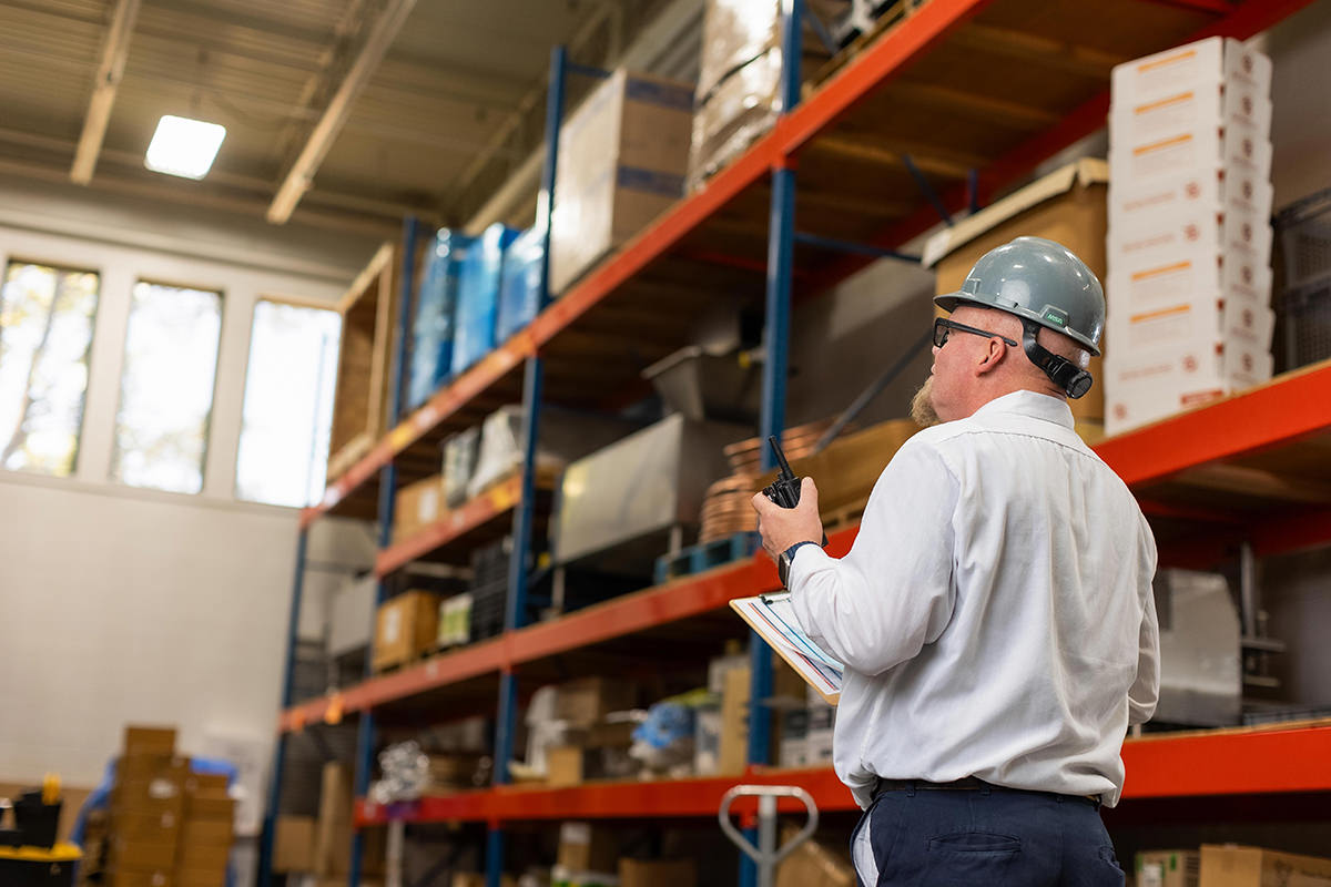 A man in a hard hat holds a clipboard and radio, overseeing shelves in a warehouse, akin to setting up group photography for an efficient snapshot of organized precision.