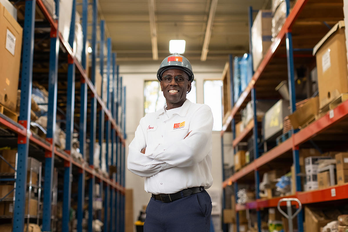 A man in a hard hat and uniform stands smiling with arms crossed in a warehouse aisle filled with shelves of boxes, as if ready to join group photography.