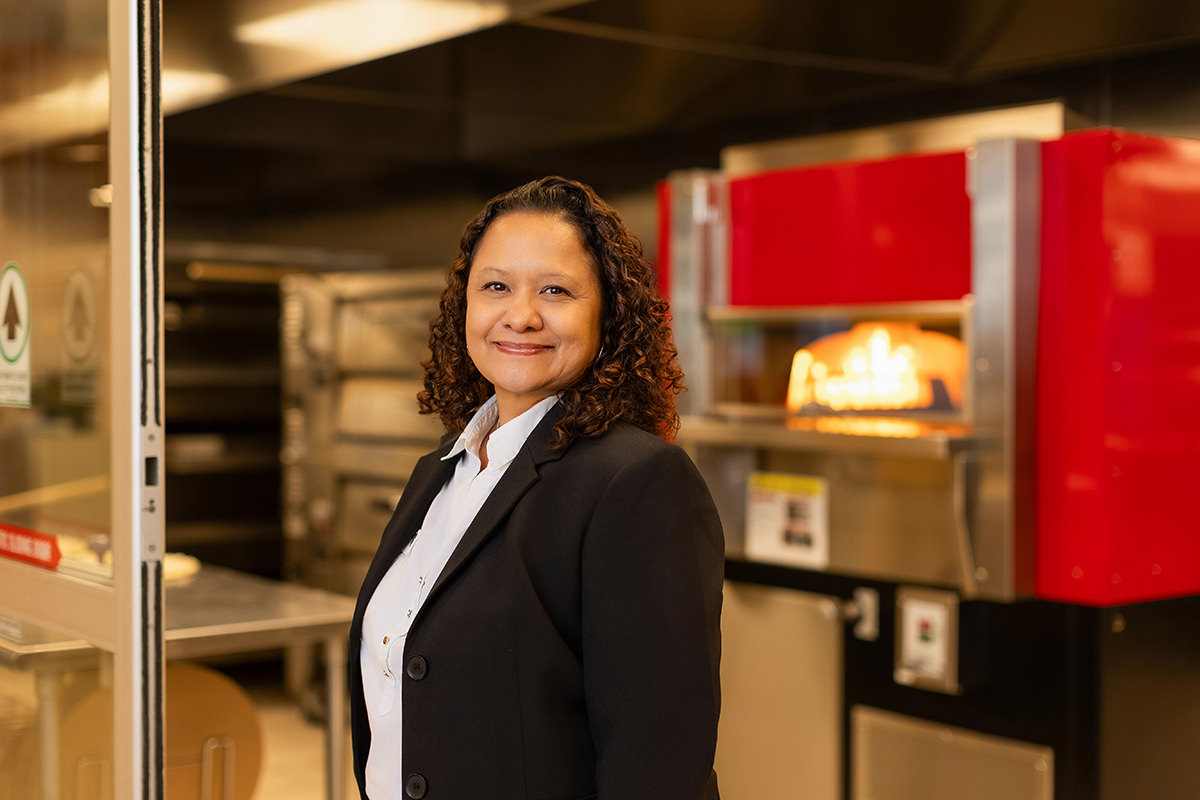 A woman in a black blazer poses confidently in a commercial kitchen, surrounded by pizza ovens, as if ready for a group photography session.