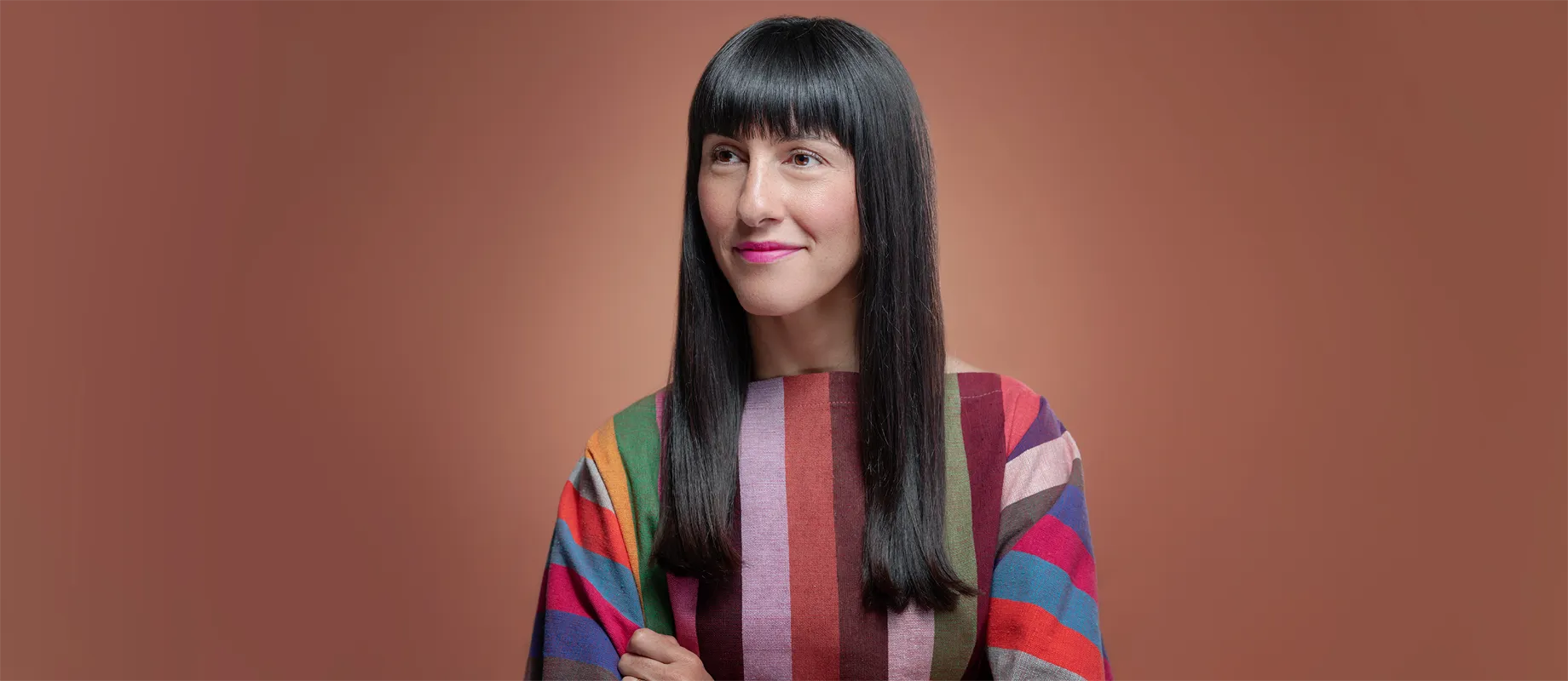 Person with long black hair and straight bangs, wearing a colorful striped top, stands against a plain brown background.