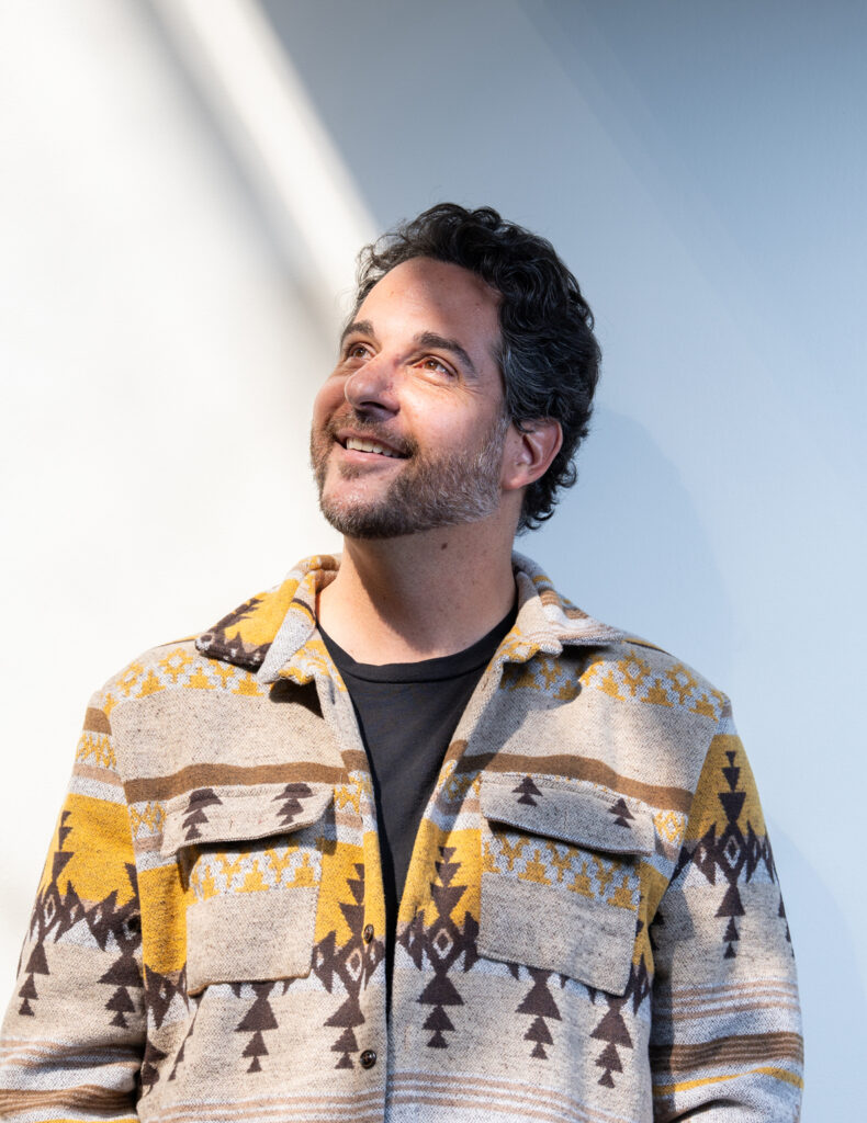 A man with dark hair and a beard, wearing a patterned jacket, smiles warmly while looking upward, captured in a professional shot by Denver Headshot Co against a light background.