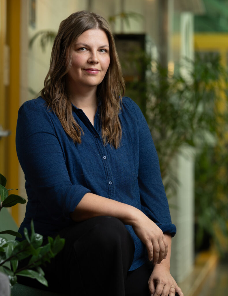 A person with long hair sits indoors, showcasing a crisp blue shirt and dark pants, framed by lush plants in the background—an ideal setting captured by Denver Headshot Co.