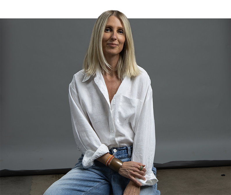 A woman with long blonde hair sits against a gray background, wearing a white shirt and blue jeans, perfect for an individual headshot.