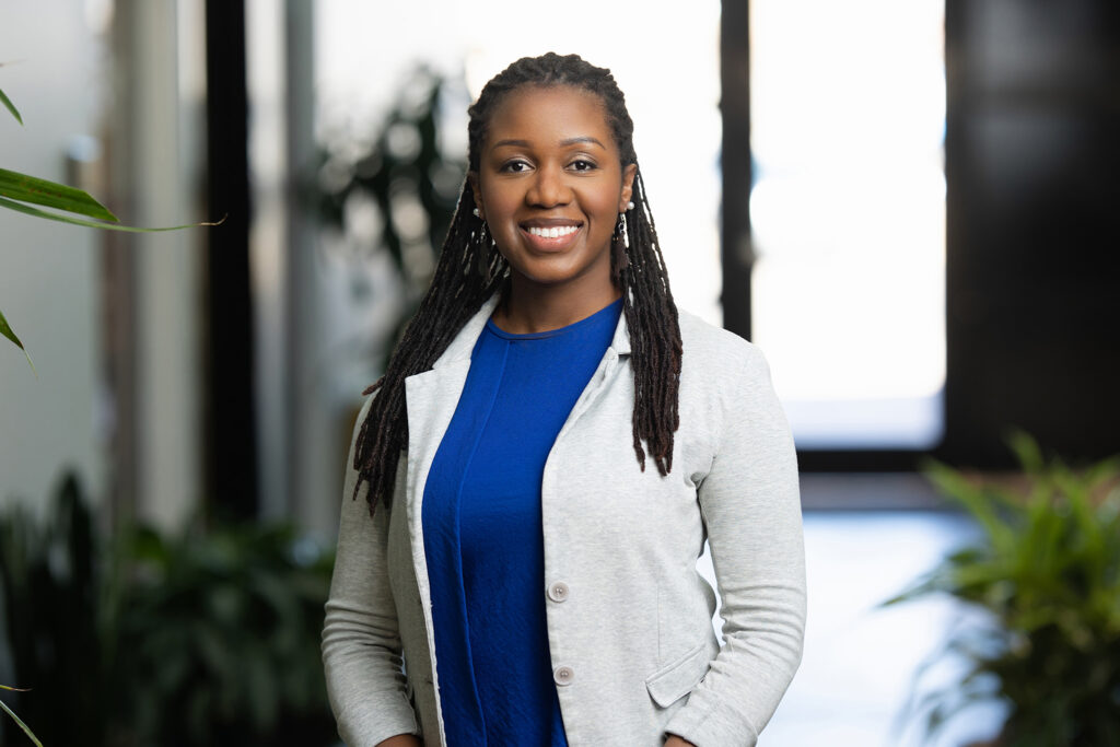 A woman in a blue dress and gray blazer stands smiling in a well-lit indoor setting with plants in the background, exuding confidence—a smart career move captured perfectly.