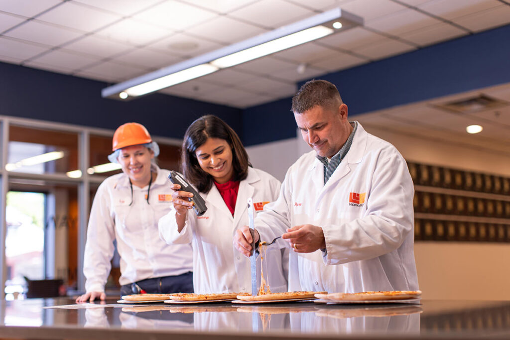 Three people in lab coats and hard hats, identified as industry leaders, examine pizzas on a table under bright lighting in a room with a tiled ceiling—a scene set for 2025 marketing's latest visual content campaign.