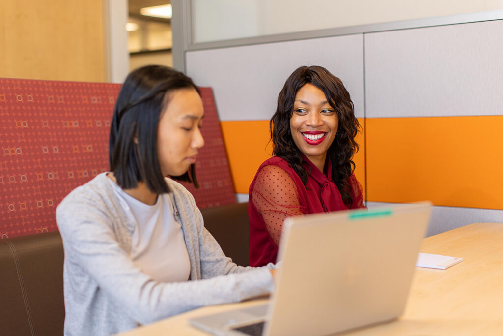 Two women sit at a table in an office setting, with red and orange dividers in the background. One types on a laptop, creating visual content for 2025 marketing strategies, while the other smiles, embodying the spirit of industry leaders shaping the future.