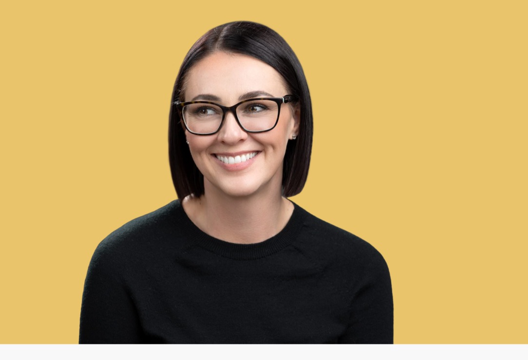 An individual with shoulder-length dark hair and glasses smiles while looking to the side against a yellow background, creating a vibrant headshot.