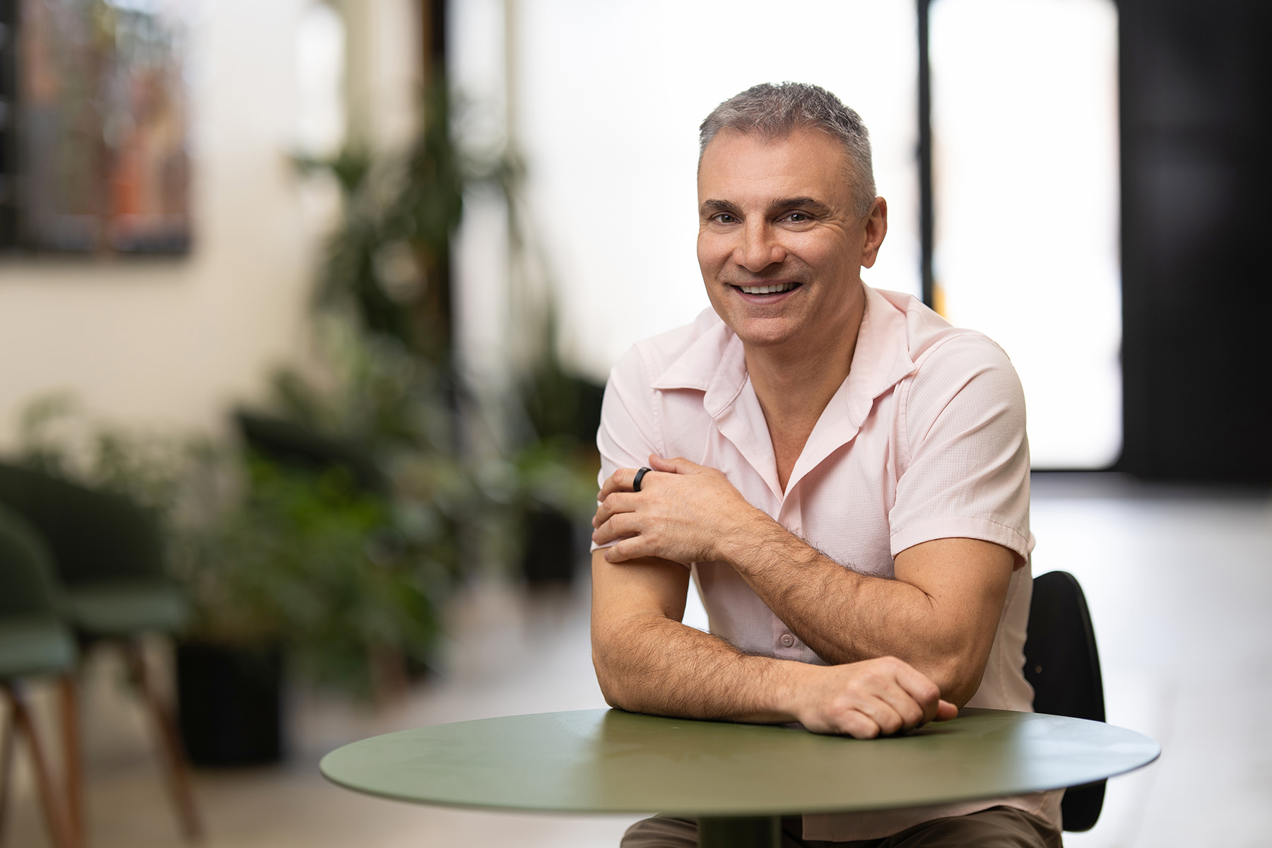 A man smiling, framed like an individual headshot, is seated at a green table in a bright room adorned with lush plants.