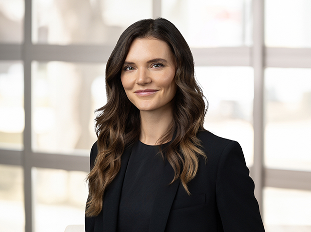 A woman with long wavy hair smiles warmly for her individual headshot, wearing a black blazer in front of a blurred window background.