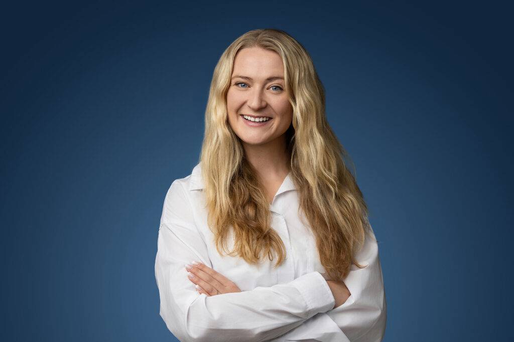 A woman with long blonde hair smiles authentically, wearing a white shirt against a blue background. In this digital world, she stands confidently with her arms crossed.