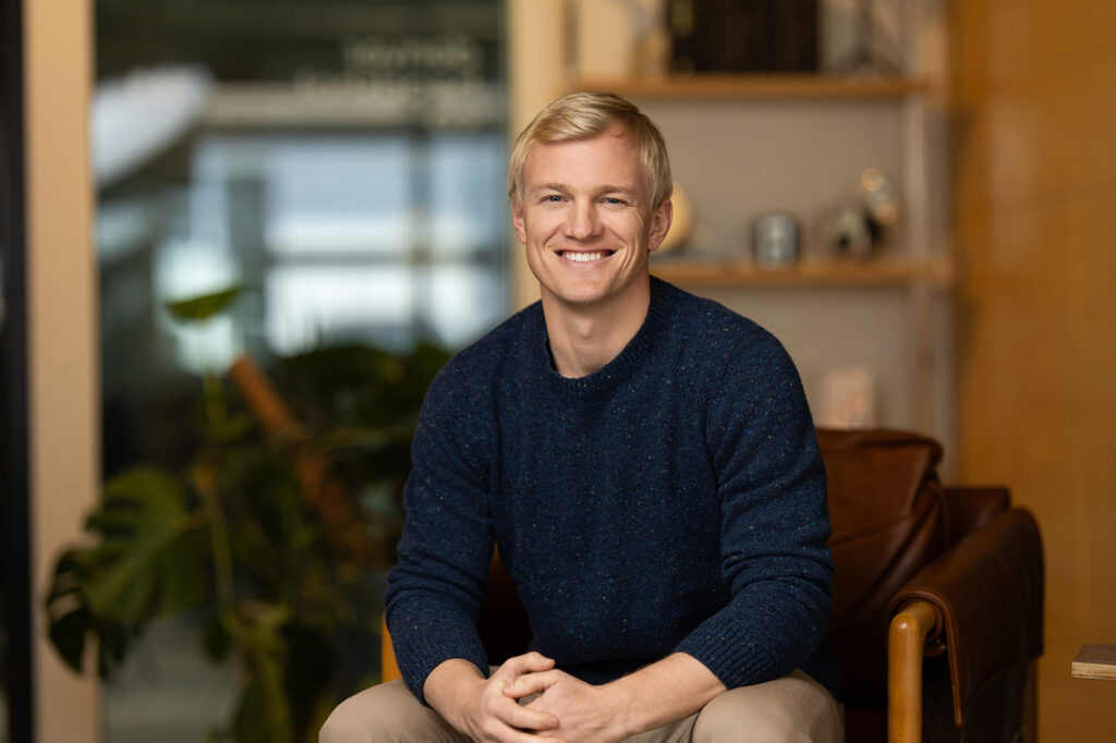 A person with short blond hair, wearing a dark sweater, is seated and smiling in a room adorned with a plant and shelves. This glimpse into their authentic space captures the power of being real amidst the digital world's influence.