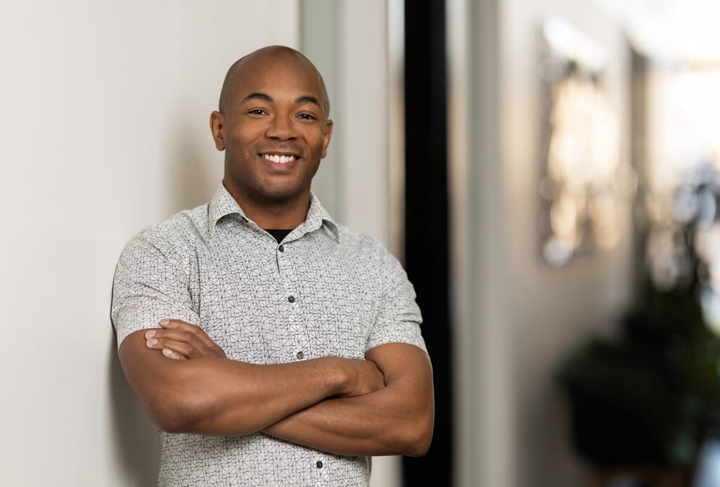 A man in a patterned shirt stands smiling with arms crossed in a bright hallway, embodying authenticity in an increasingly digital world.