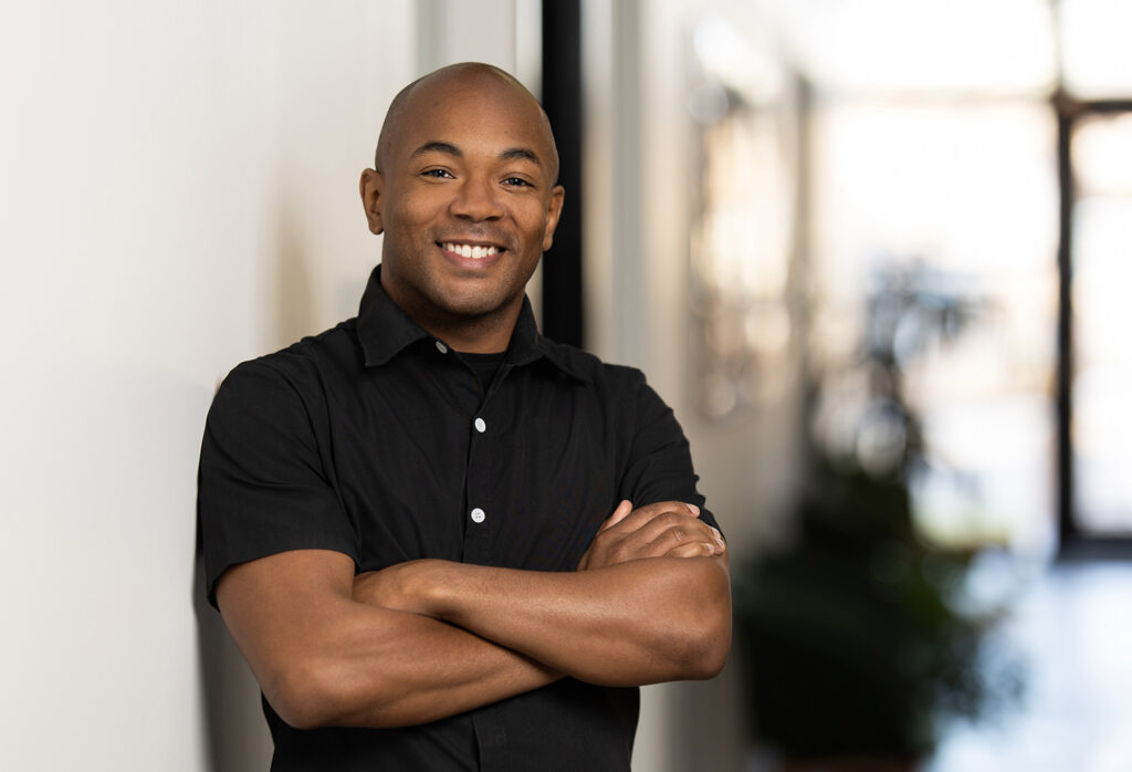 A man in a black shirt stands with arms crossed, leaning against a white wall. He is smiling in an authentically warm way, embodying realness in an otherwise digital world. The background is softly lit, featuring a blurry interior setting.