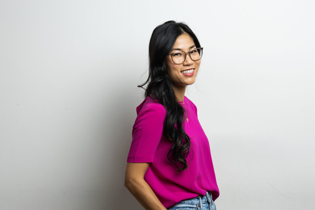 A person with long dark hair, wearing glasses and a bright pink shirt, embodies authenticity while smiling against a plain white background.